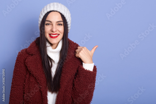 Close up portrait of brunette woman with long dark hair and red pomade, female wearing maroon faux fur coat and white winter cap, pointing aside with her thumb, looking at camera. Copy space. photo