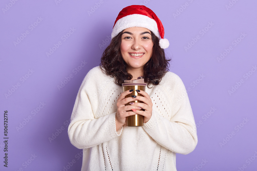 Closeup portrait of magnificent beautiful brunette looking directly at camera, smiling sincerely, wearing white sweater and red santa claus hat, holding thermal mug in both hands. Winter concept. Closeup portrait of magnificent beautiful brunette looking directly at camera, smiling sincerely, wearing white sweater and red santa claus hat, holding thermal mug in both hands. Winter concept.