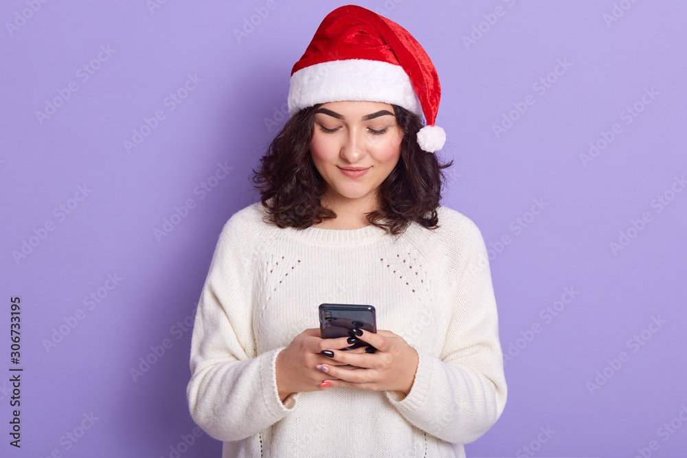 Image of pleasant cute dark haired young lady having peaceful facial expression, holding mobile phone in both hands, chatting, using social networking sites. People and new technologies concept. Image of pleasant cute dark haired young lady having peaceful facial expression, holding mobile phone in both hands, chatting, using social networking sites. People and new technologies concept.