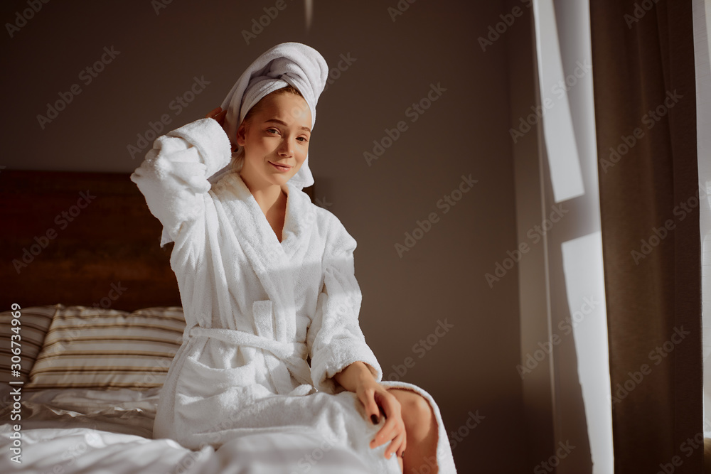 Nice attractive girl with natural face, looking at camera with pleasant expression, wearing bathrobe and white towel on head, sitting on bed in front of window with curtains, home time concept Nice attractive girl with natural face, looking at camera with pleasant expression, wearing bathrobe and white towel on head, sitting on bed in front of window with curtains, home time concept