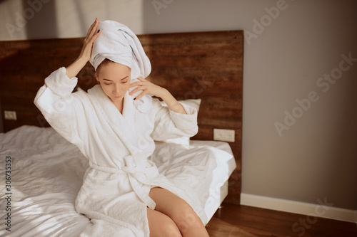 Cheerful female model enjoying time alone in bedroom after shower, dressed in white bathrobe, keeping hands on towel, looking away, healthy lifestyle, relax, softness concept