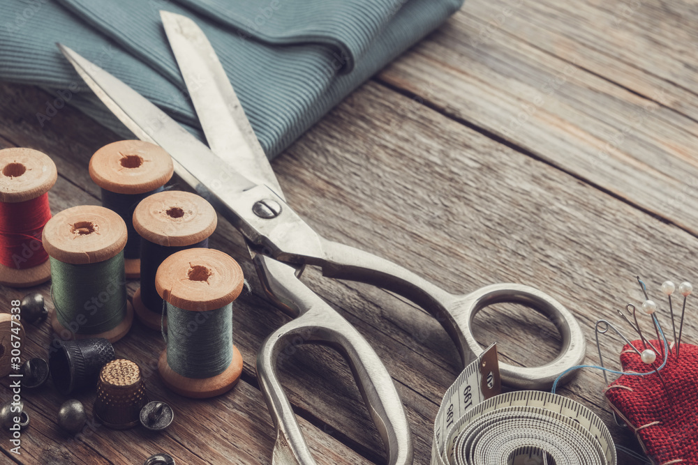 Retro sewing items: tailoring scissors, cutting knife, thimble, wooden thread spools, cushion for including pins, fabrics and sewing accessories. - obrazy, fototapety, plakaty 