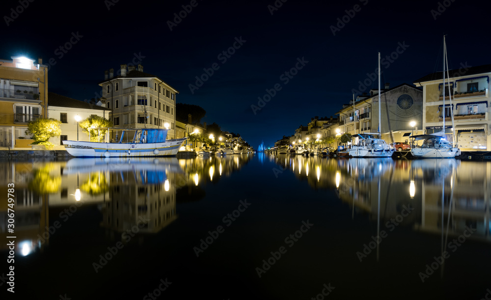 Nighttime shot of Port of Grado in Italy mirroring in water, with passageway to adriatic sea and thawed boats