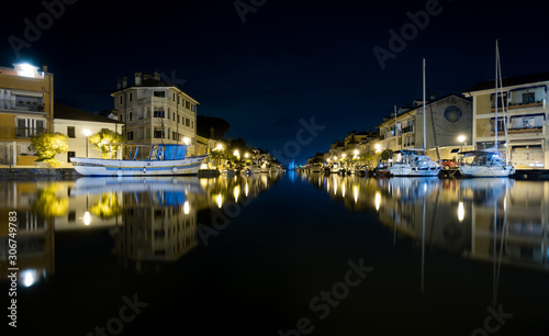 Nighttime shot of Port of Grado in Italy mirroring in water, with passageway to adriatic sea and thawed boats