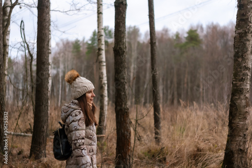 portrait in profile of beautiful young female looking in to the distanse. Girl walks in the frosty fresh air in the forest. She is wearing a gray jacket and knitted hat with a pompom.