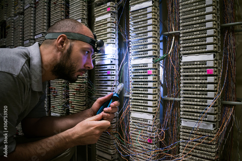 A technician with a flashlight on his head works in a dark server room. The specialist fixes the problem of communication. Man cuts a telephone wire near a patch cross panel.