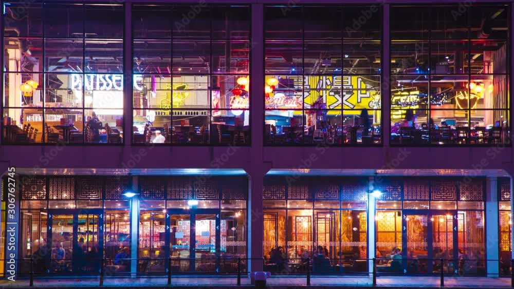 Motion blur time lapse of people passing & dining in busy restaurant interior at night with glowing neon signs