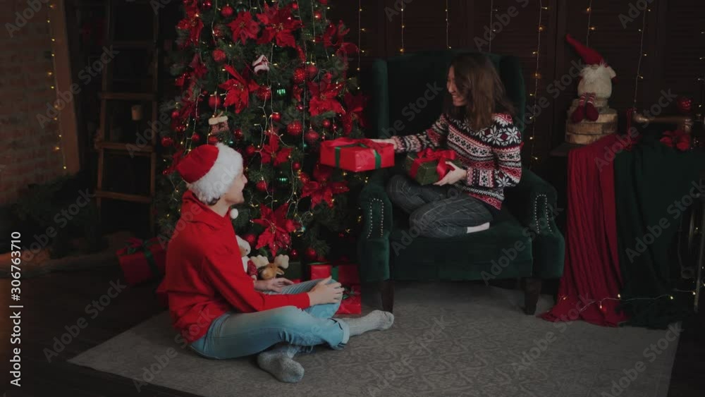 A funny guy and an Asian girl are sitting next to the Christmas Tree. Young couple give each other New Year's gifts. Family celebrates New Year at home. Racial Relations at Christmas. A funny guy and an Asian girl are sitting next to the Christmas Tree. Young couple give each other New Year's gifts. Family celebrates New Year at home. Racial Relations at Christmas.
