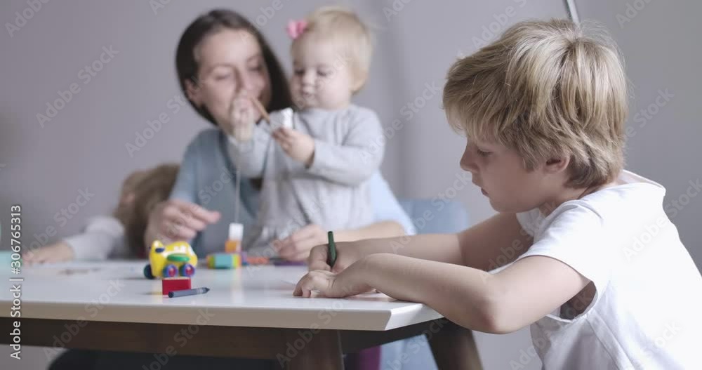 Close-up side view of blond Caucasian schoolboy in white T-shirt drawing. His mother and two sisters sitting at the background and playing. Happy calm day in big family. Cinema 4k ProRes HQ. Close-up side view of blond Caucasian schoolboy in white T-shirt drawing. His mother and two sisters sitting at the background and playing. Happy calm day in big family. Cinema 4k ProRes HQ.