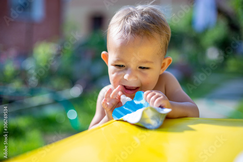 cute little baby put his finger in the package of fruit puree in pouch  in front of the yellow table. on the background is a green garden on a sunny day in blur, close up