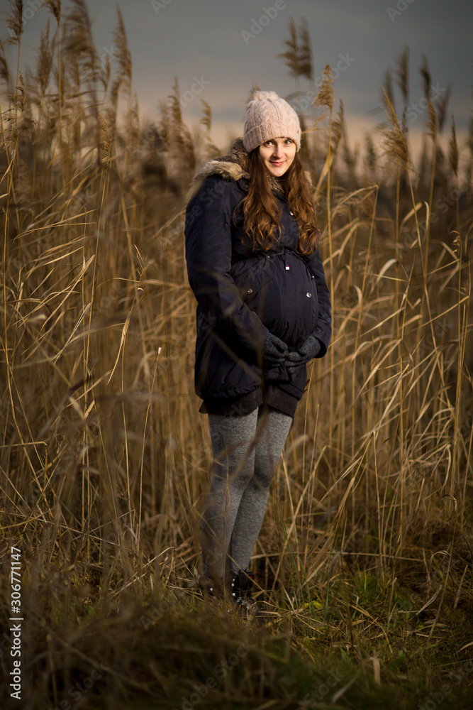 Pregnant woman in a forest or grass in autumn or winter. Pregnancy photo of standing woman in a high grass. wearing hat jacket cold whether Pregnant woman in a forest or grass in autumn or winter. Pregnancy photo of standing woman in a high grass. wearing hat jacket cold whether