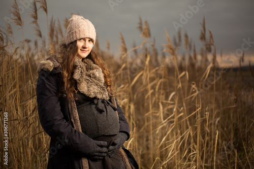 Pregnant woman in a forest or grass in autumn or winter. Pregnancy photo of standing woman in a high grass. wearing hat jacket cold whether