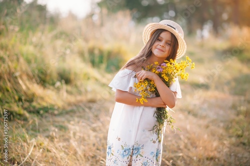Outdoor shot of pleasant looking young girl with tanned healthy skin  dressed in white dress and summer hat  poses in park with confident satisfied expression  likes recreation. Beautiful young woman