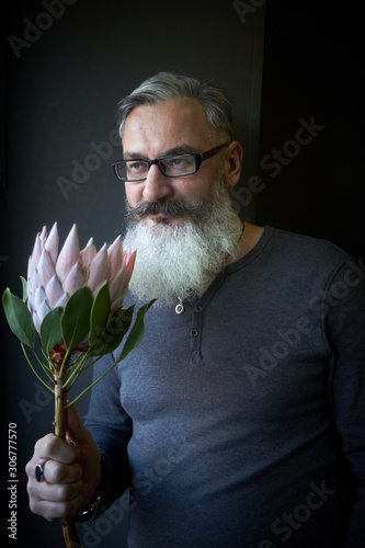 Gray-haired man with glasses and a beard holds a pink protea in his hands, selective focus