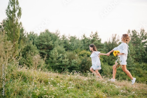 Cheerful mother and her little daughter having fun together in the summer background. Happy family in the nature background. Cute girls with colorful flowers.