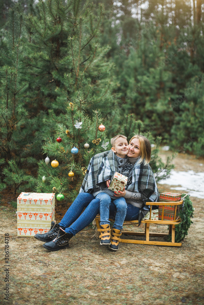 pretty young mother with child son while sitting on the wooden decorated sledge outdoors in winter forest, covered with checkered plaid together. Family winter holidays outdoors.