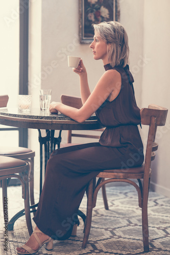 Toned picture of side view of beautiful blonde lady sitting in cafe or restaurant near window and drinking coffee