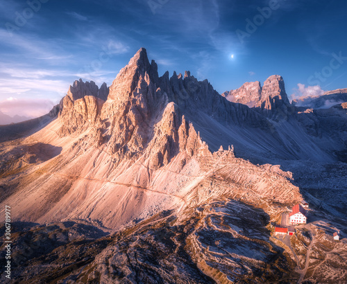 Mountains with beautiful house and church at sunset in autumn. Landscape with buildings, high rocks with trails, blue sky with moon and clouds at dusk. Aerial view of Tre Cime park in Dolomites, Italy