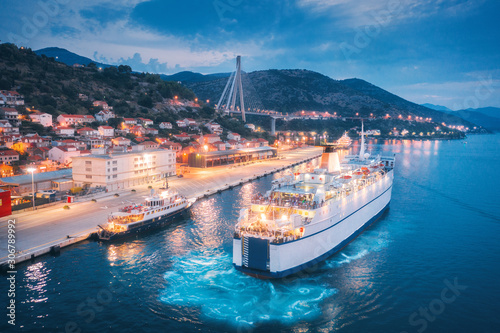 Aerial view of cruise ship in port at night. Landscape with ships and boats in harbour, city lights, buildings, mountains, blue sea at sunset. Top view. Luxury cruise. Floating liner at harbor in dusk photo