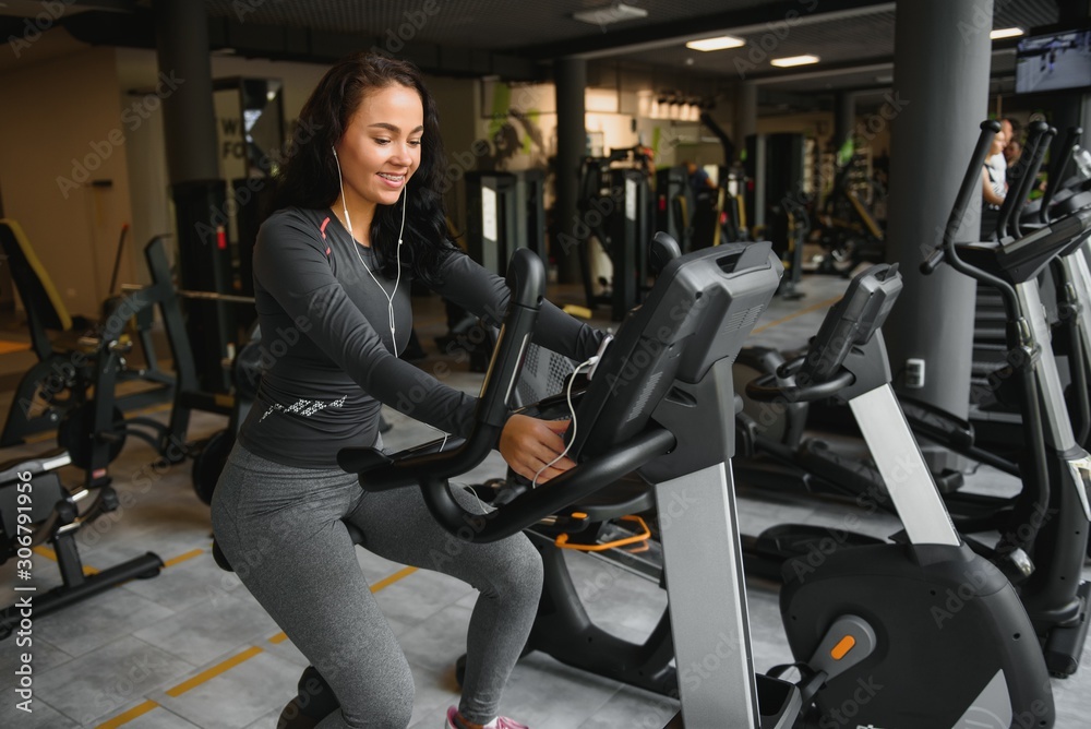 Young woman with headphones doing exercises on stationery bicycle in a gym or fitness center. Young sporty woman in gym listen music from smartphone. Women doing cardio exercises.