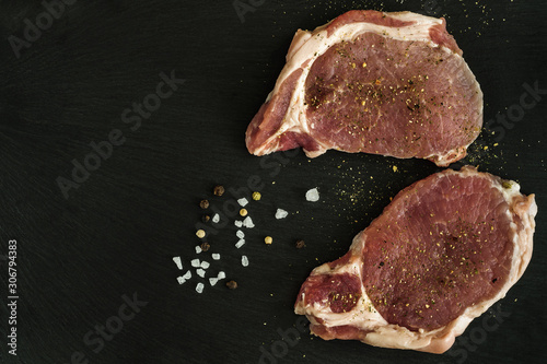 two raw pork steaks with spices on a black stone board close-up. top view with copy space