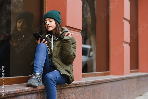 young stylish beautiful teen girl listening to music, mobile phone, headphones, enjoying, denim outfit, smiling, happy, cool accessories, having fun, laughing, park