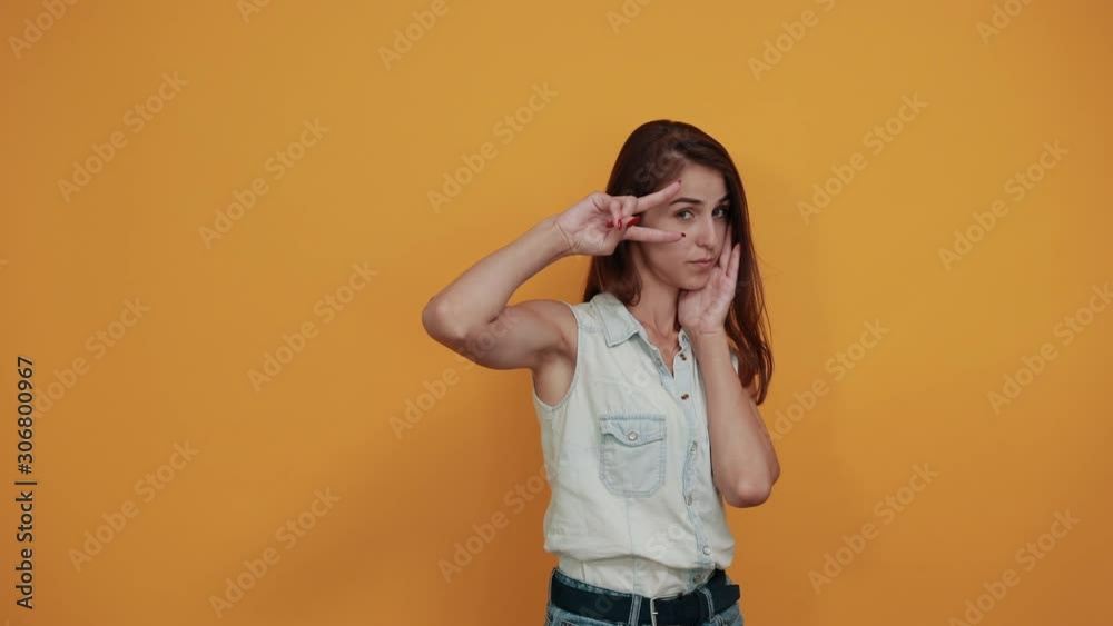 Boring caucasian young woman in blue denim shirt doing victory gesture, keeping hand on cheek, sleep isolated on orange background in studio. People sincere emotions, lifestyle concept. Boring caucasian young woman in blue denim shirt doing victory gesture, keeping hand on cheek, sleep isolated on orange background in studio. People sincere emotions, lifestyle concept.