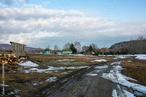 Russian village. Bad dirt road in the middle of a Russian village on the background of wooden houses and a fence. Dirty rural road in the spring in puddles.