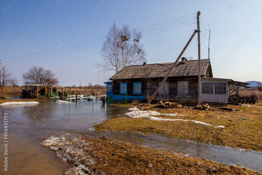 Russian village. An old rustic wooden house behind a low wooden picket fence in spring. Spring puddles on the background of a village hut in Russia. Russian village. An old rustic wooden house behind a low wooden picket fence in spring. Spring puddles on the background of a village hut in Russia.