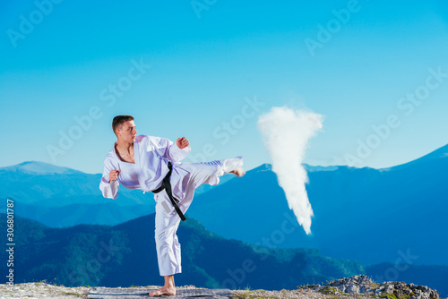 Fit karate athlete kicking a cup filled whit fluor causing a big splash while wearing a white kimono on top of a mountain on a sunny day.