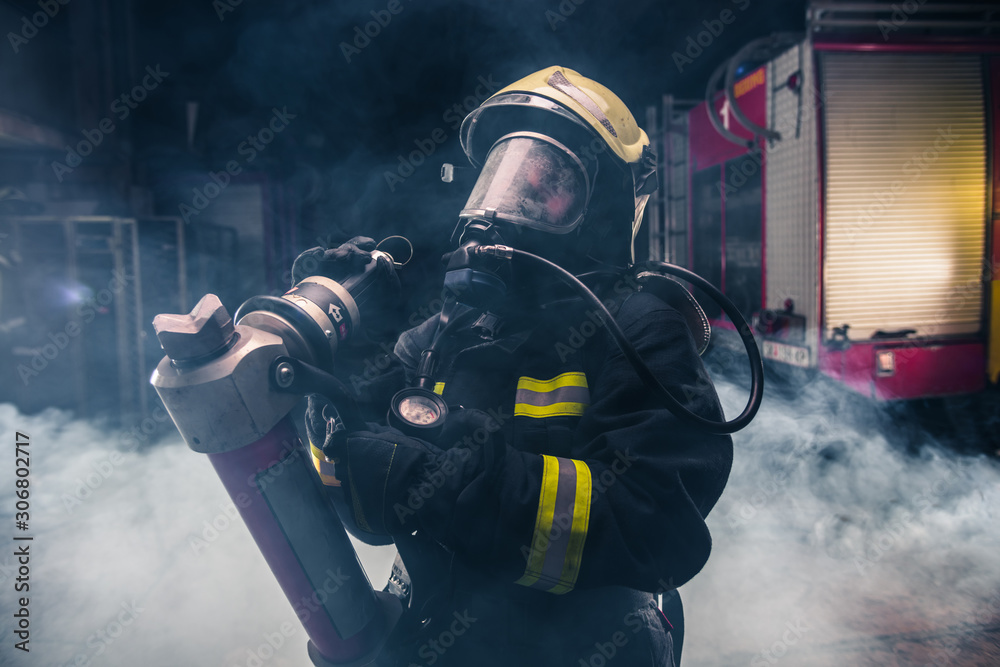 Portrait of a female firefighter wearing a helmet and all safety equipment a while holding a tomahawk and wearing an oxygen mask indoors surrounded by smoke