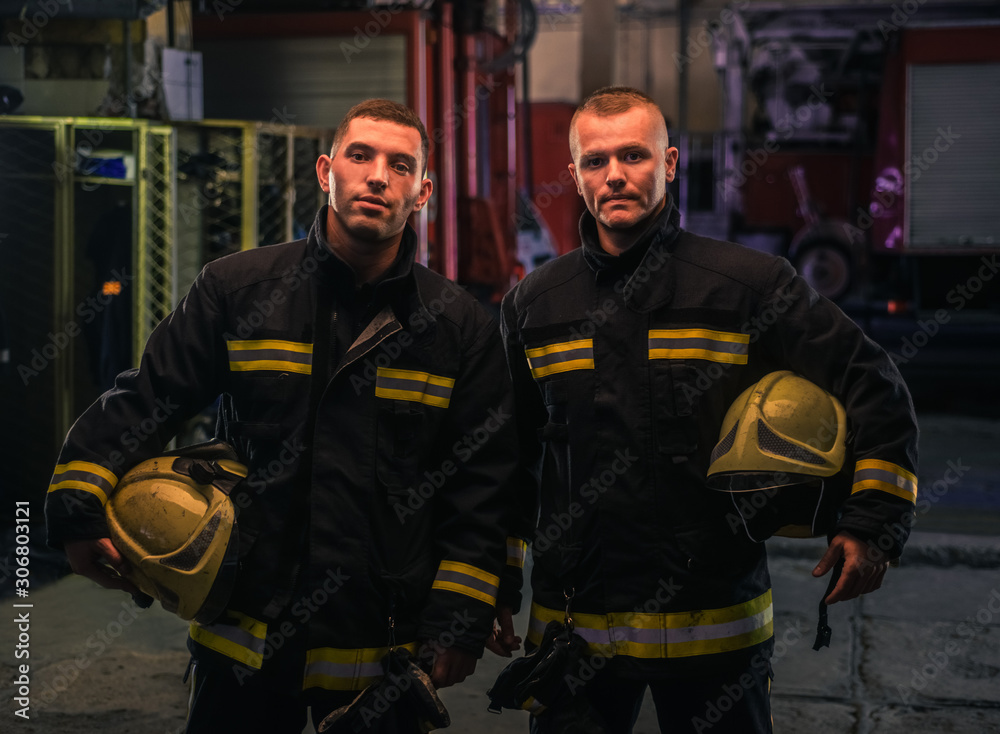 Fototapeta premium Portrait of two young firemen in uniform standing inside the fire station .