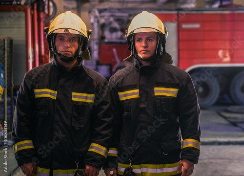 Portrait of two young firemen in uniform standing inside the fire station .