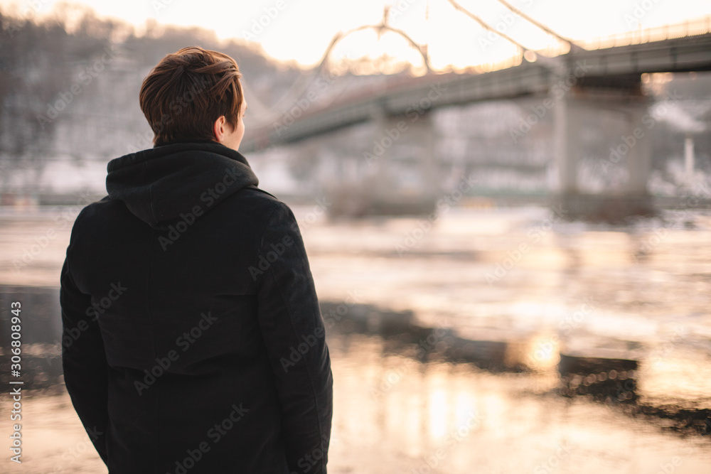 Back view of thoughtful young man looking at river while standing outdoors in winter - obrazy, fototapety, plakaty 