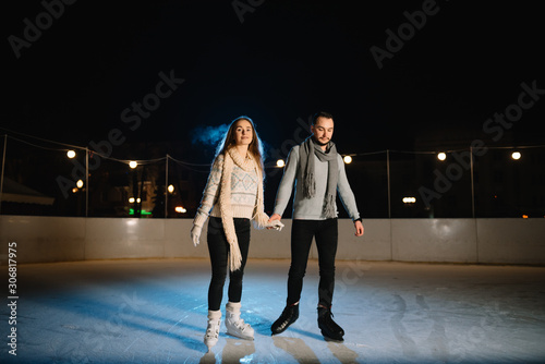 Man and woman young family Happy smiles hold hands skate on winter rink at night, with bokeh lights.
