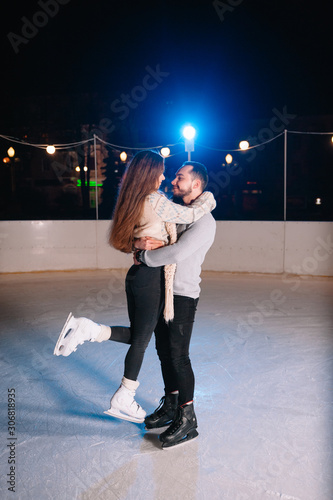 Couple on the city rink in a winter evening. Guy helping nice girl to skate on the ice in the dark night and twinkles lighting above them