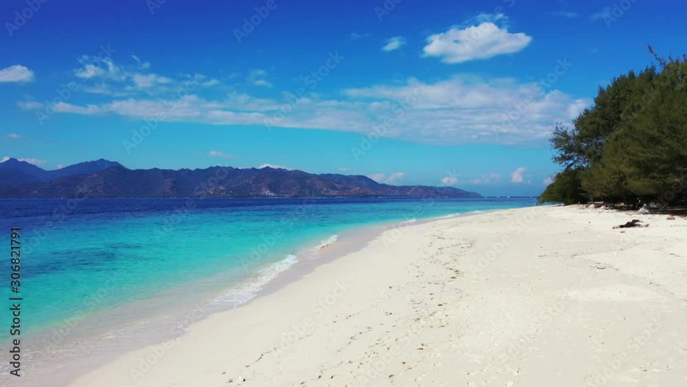 Pristine white beach with fine sand washed by white waves of blue azure sea, bordered by high mountains on a bright blue sky with frozen clouds in Bali Pristine white beach with fine sand washed by white waves of blue azure sea, bordered by high mountains on a bright blue sky with frozen clouds in Bali