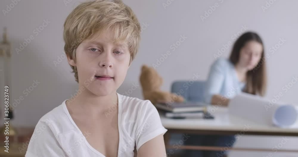 Portrait of tired blond Caucasian boy with grey eyes yawning and looking at camera. His young mother sitting at the table at the background. Guy in white T-shirt posing at home. Cinema 4k ProRes HQ.