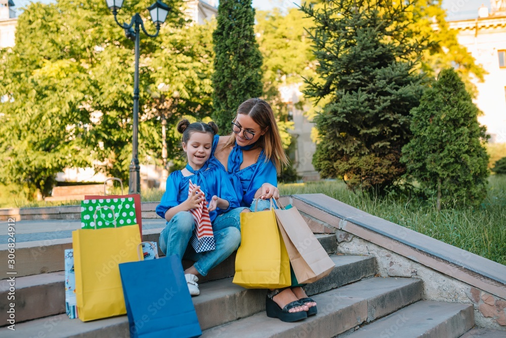 Beautiful mom and her cute little daughter are holding shopping bags, looking at camera and smiling while standing outdoors. Shopping concept. Beautiful mom and her cute little daughter are holding shopping bags, looking at camera and smiling while standing outdoors. Shopping concept.