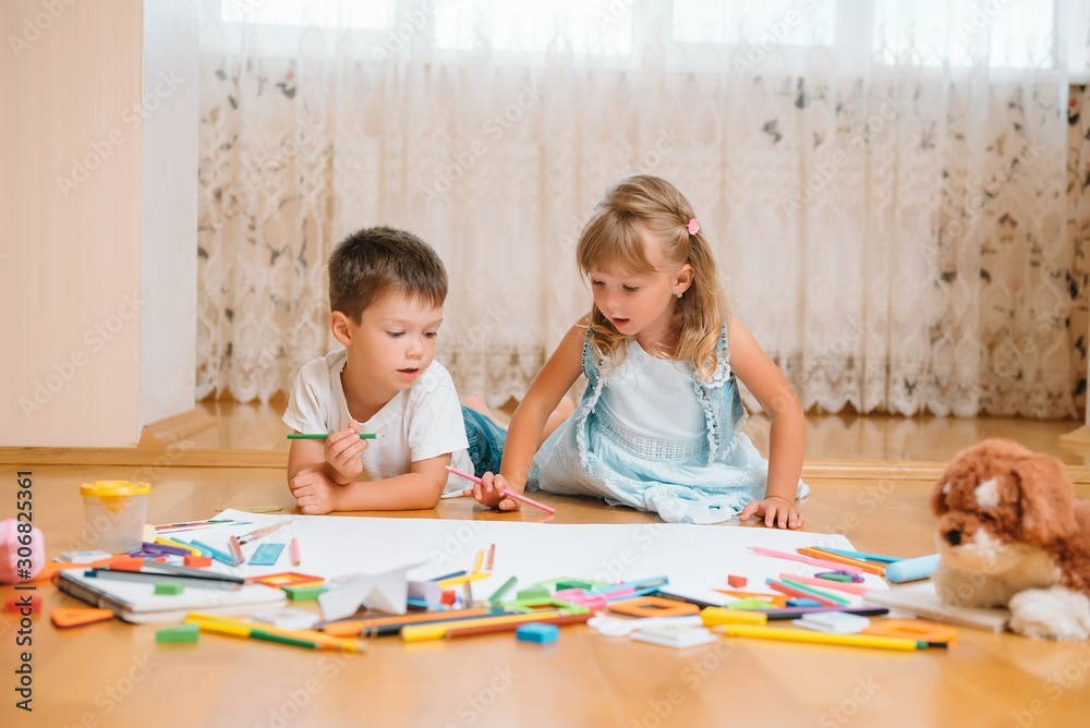 Kids drawing on floor on paper. Preschool boy and girl play on floor with educational toys - blocks, train, railroad, plane. Toys for preschool and kindergarten. Children at home or daycare.