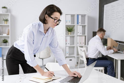 Confident businesswoman bending over desk in front of laptop while networking