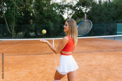 Portrait of fashionable woman in red and white clothing with tennis racket posing at tennis net on court. Sports Fashion