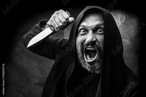 Black and white portrait of a bald bearded man in a hood on a dirty gray background. Maniac with a knife concept.