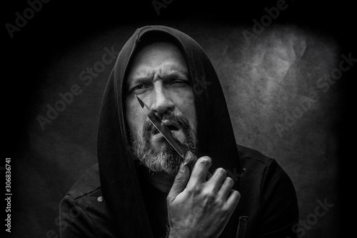 Black and white portrait of a bald bearded man in a hood on a dirty gray background. Maniac with a knife concept.