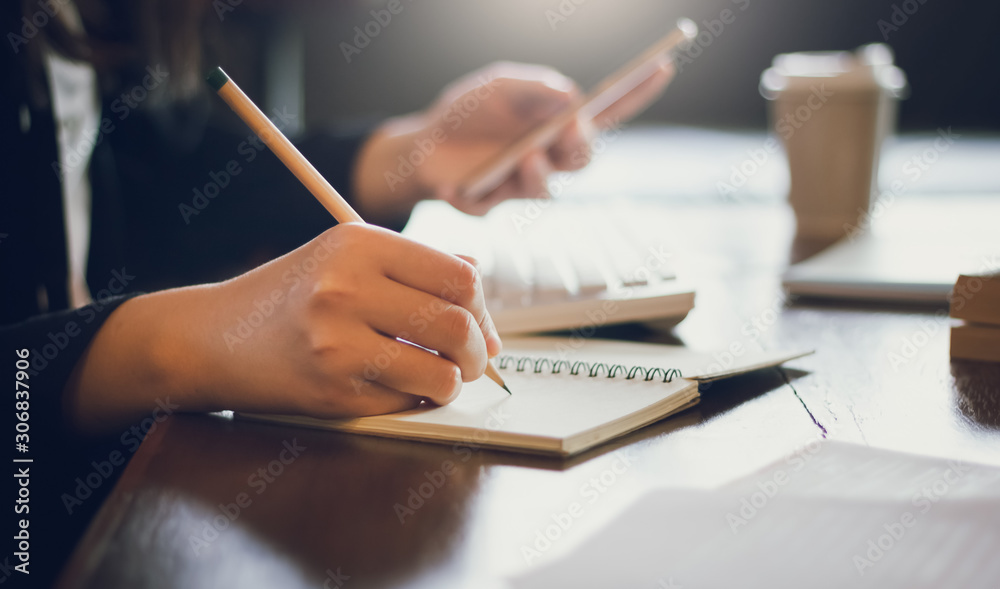 Close-up of hand young businesswoman writing information on notebook and using mobile phone working with computer