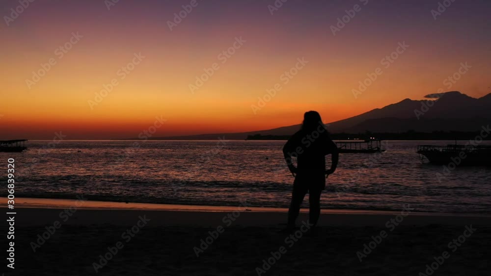Silhouette of girl standing on exotic beach in front of dark sea surface reflecting a low light of twilight orange sky over Indonesian horizon Silhouette of girl standing on exotic beach in front of dark sea surface reflecting a low light of twilight orange sky over Indonesian horizon