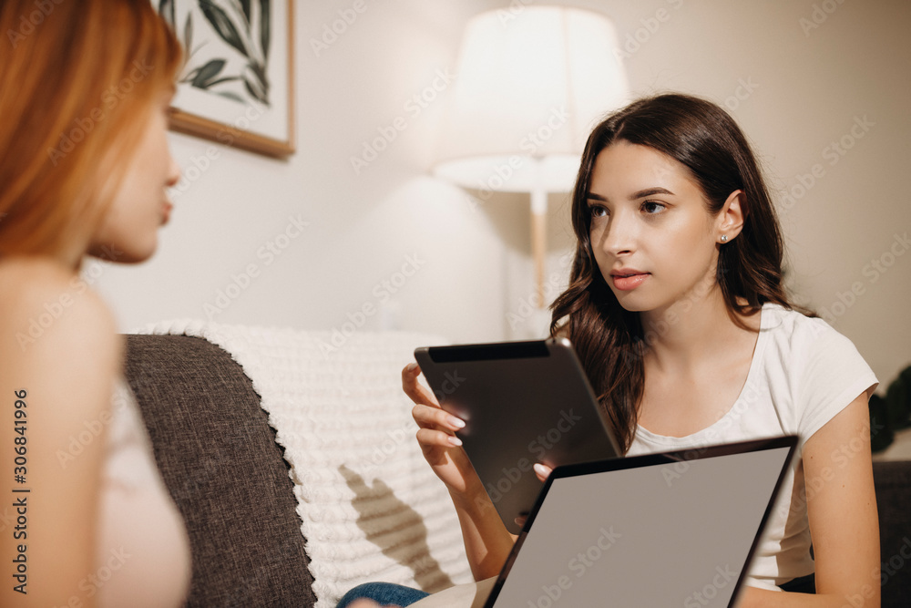 Two beautiful girlfriend sitting face to face talking and storytelling while holding tablet and laptop on their hands sitting on the couch. Two beautiful girlfriend sitting face to face talking and storytelling while holding tablet and laptop on their hands sitting on the couch.