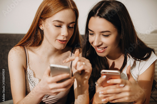 Close up portrait of a lovely young woman with red hair and freckles showing on her smartphone screen to her female friend while sitting on the couch.