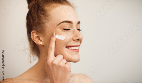 Fotografia Close up of a young female with red hair and freckles applying white cream on her face laughing with closed eyes isolated on white background