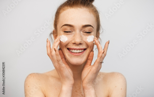 Close up of a beautiful woman with red hair and freckles playing with both hand cream on her face laughing with closed eyes against a white wall.